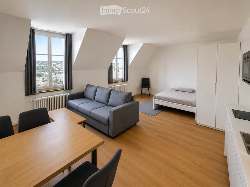 A modern, compact studio apartment with wooden flooring, large windows offering a city view, a grey sofa, a wooden dining table with chairs, a single bed, and a small kitchen area with a microwave.