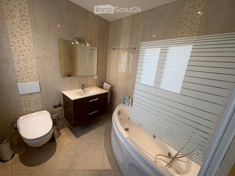 Modern bathroom with beige tiles, a toilet, a sink with a dark wooden cabinet, a bathtub with a glass shower enclosure, and a large mirror with built-in lighting.