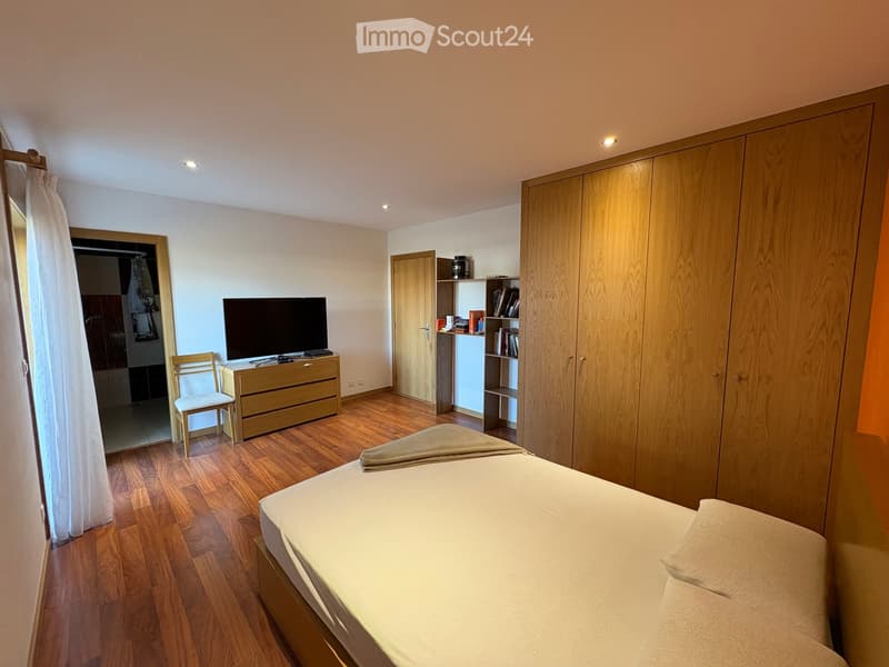 Well-lit bedroom with wooden flooring, a neatly made bed, a wooden wardrobe, a dresser with a TV, a chair, and a shelf with books and decorative items.