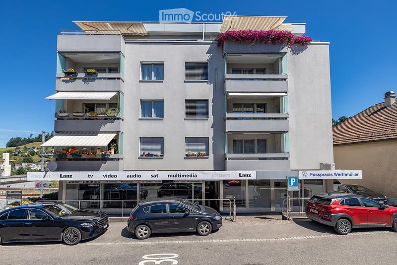 Modern multi-story building with balconies, awnings, and storefronts. Several cars are parked in front.