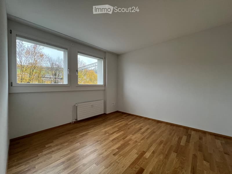Empty room with wooden flooring, white walls, and large windows offering a view of trees and a bridge outside.