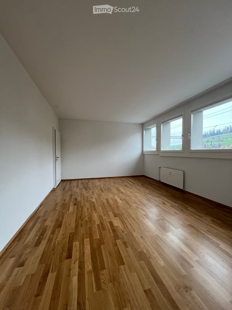 Empty room with wooden flooring, white walls, and large windows offering a view of the outdoors.