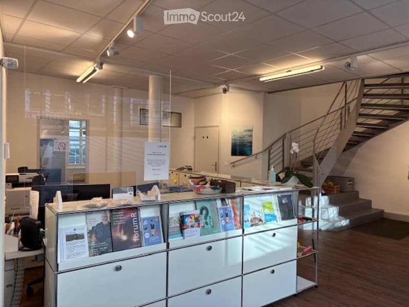 Office space with a glass wall, a staircase, a desk, a monitor, a cabinet, and a shelf with books.