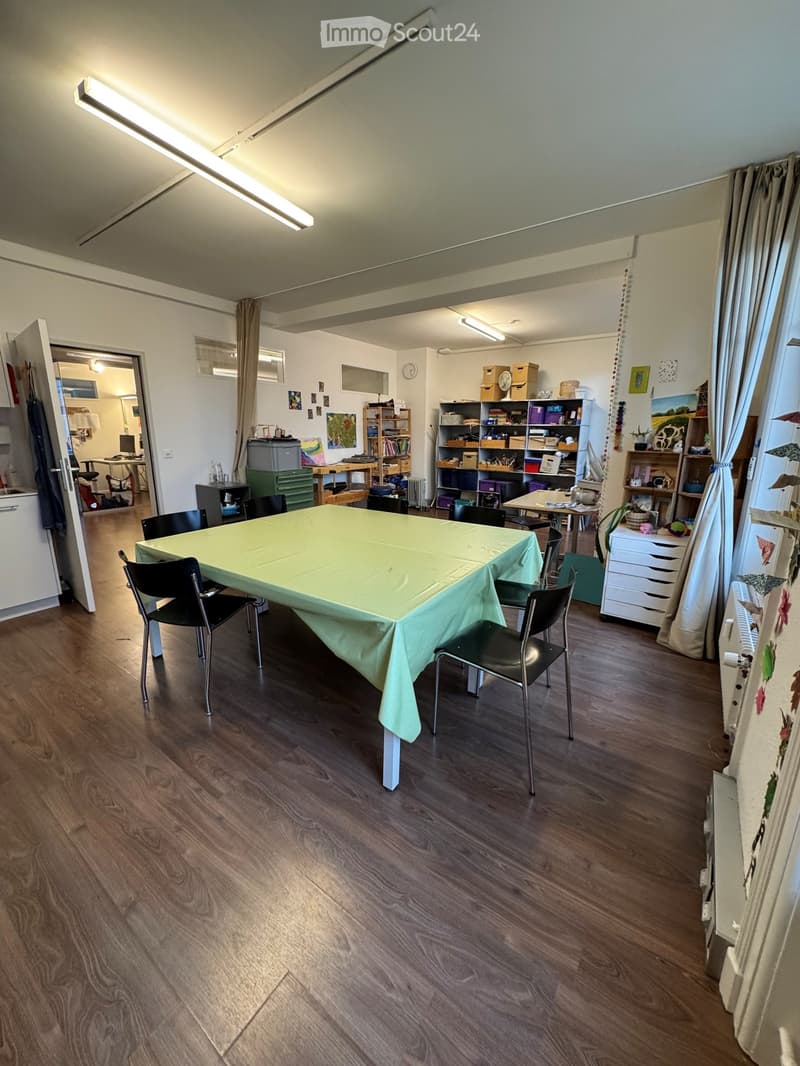 large room with wooden floor, long table with green cloth, black chairs, shelves with various items, radiator, ceiling lights