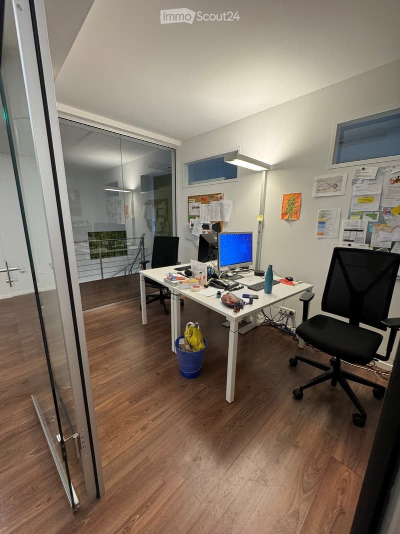 An office with a desk, two chairs, a monitor, a lamp, a trash bin, and a bag on the floor.