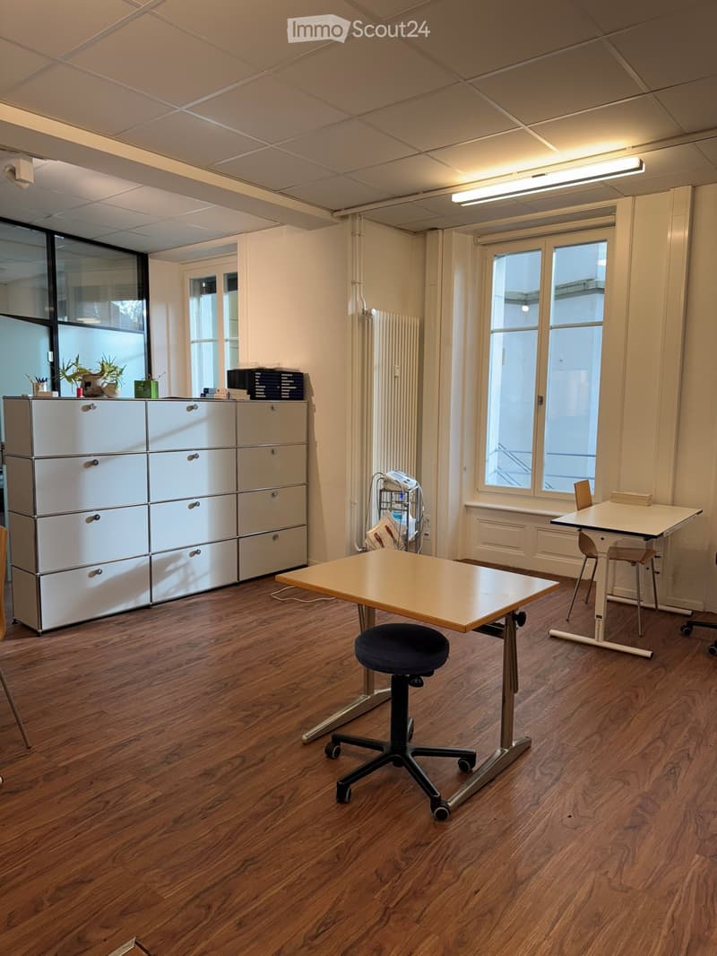 empty room with wooden floor, white walls, large windows, two desks, one with a chair, one with a stool, a white cabinet, a radiator, and a ceiling light