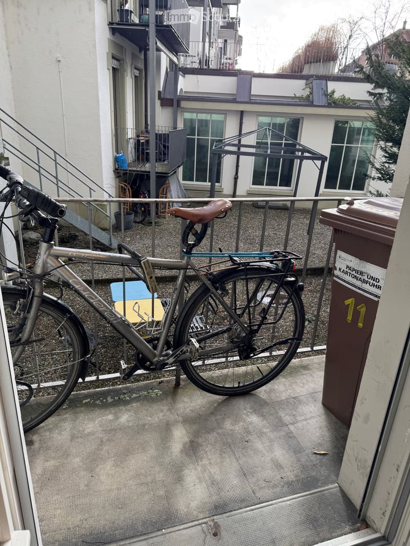A bicycle is parked on a balcony with a metal railing, a trash bin, and a blue chair.