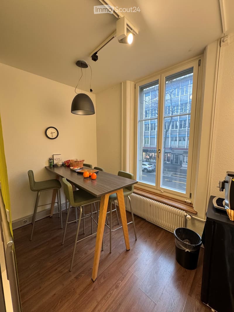 Dining area with a wooden table, green chairs, a radiator, a clock, and a window with a view of the street outside.
