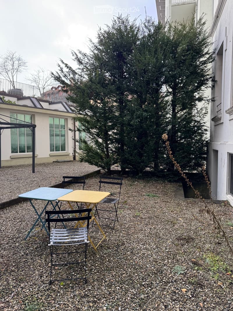 A graveled garden with a canopy, tables, and chairs, surrounded by trees and buildings.