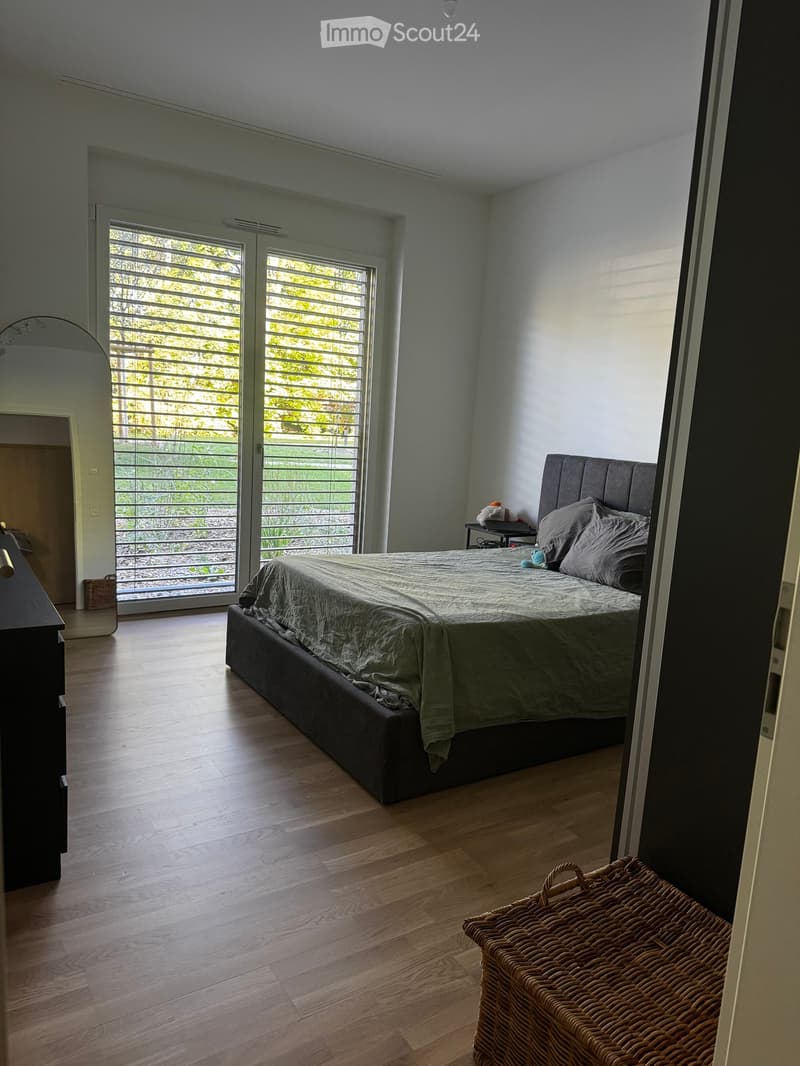 Modern bedroom with wooden flooring, large bed with grey bedding, nightstand, mirror, and wicker basket near the door.