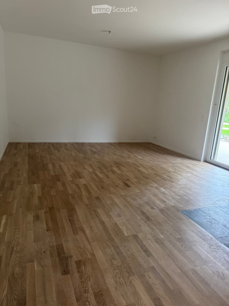 Empty room with wooden flooring and white walls, featuring a large window with a view of greenery.