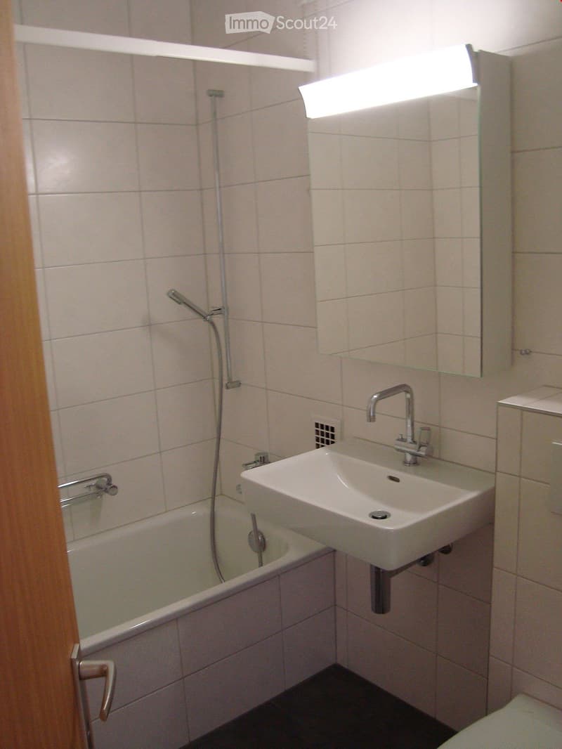 Modern bathroom with a bathtub, sink, and mirror. Light-colored tiles cover the walls and floor, and a showerhead is mounted on the wall.