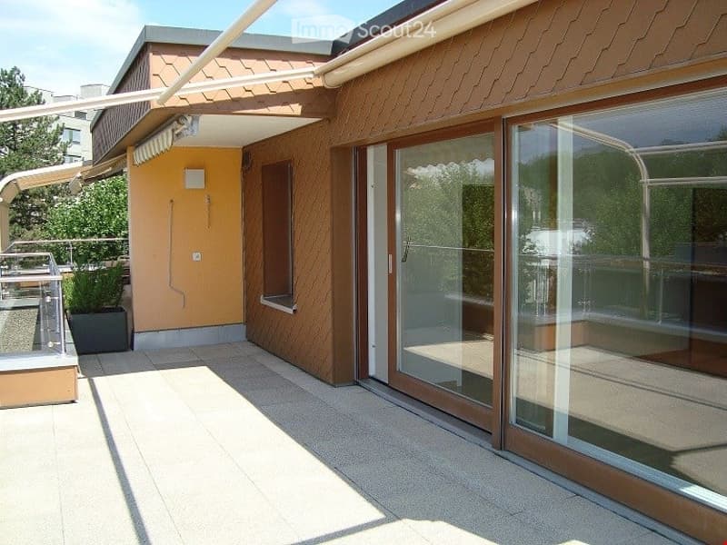 Modern balcony with large glass sliding doors, wooden decking, and a small potted plant. The balcony is covered by a retractable awning and has a clean, minimalist design.