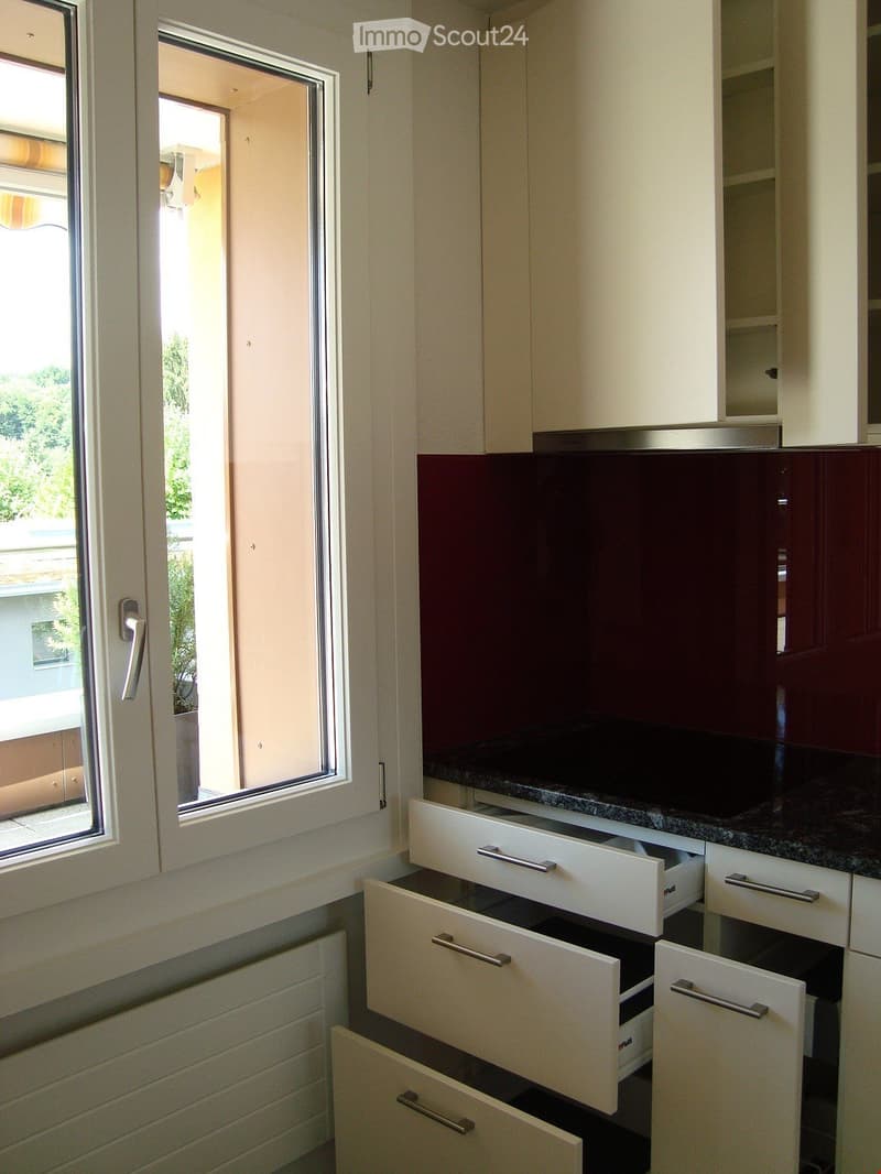 Modern kitchen with white cabinets, open drawers, and a dark countertop, featuring a large window with a view of greenery outside.