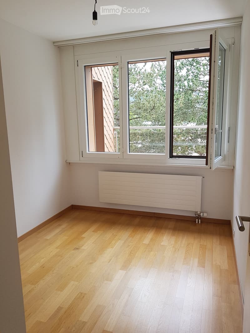 Empty room with light wooden flooring, white walls, and large windows offering a view of greenery. A radiator is mounted on the wall below the windows.