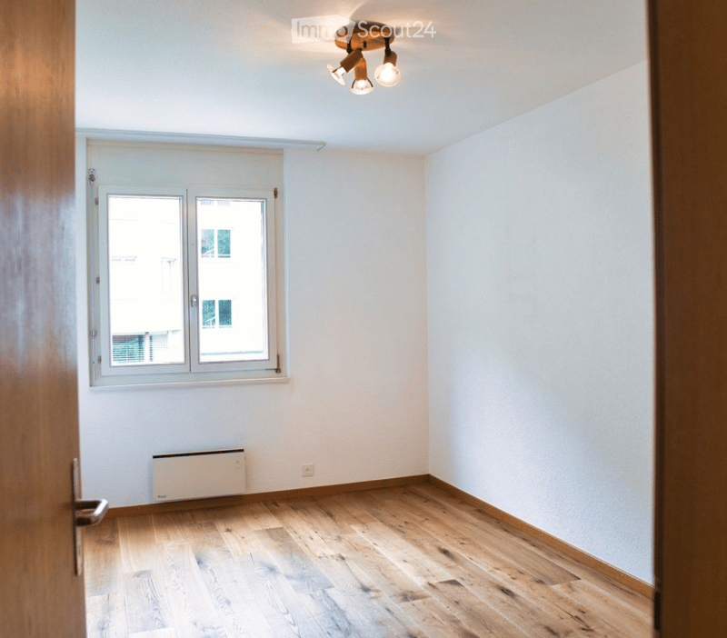 Empty room with wooden flooring, white walls, a window with a view, and a radiator under the window.