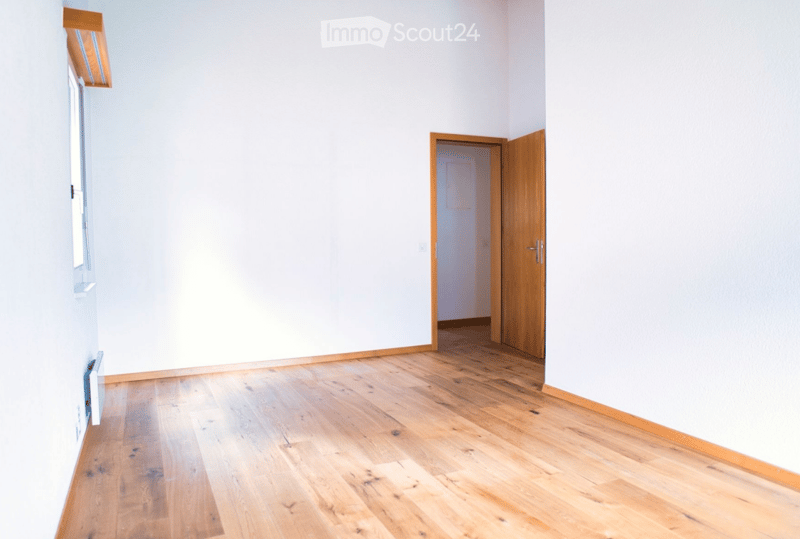 Empty room with light wooden flooring and white walls, featuring a wooden door and a window with a built-in shelf.