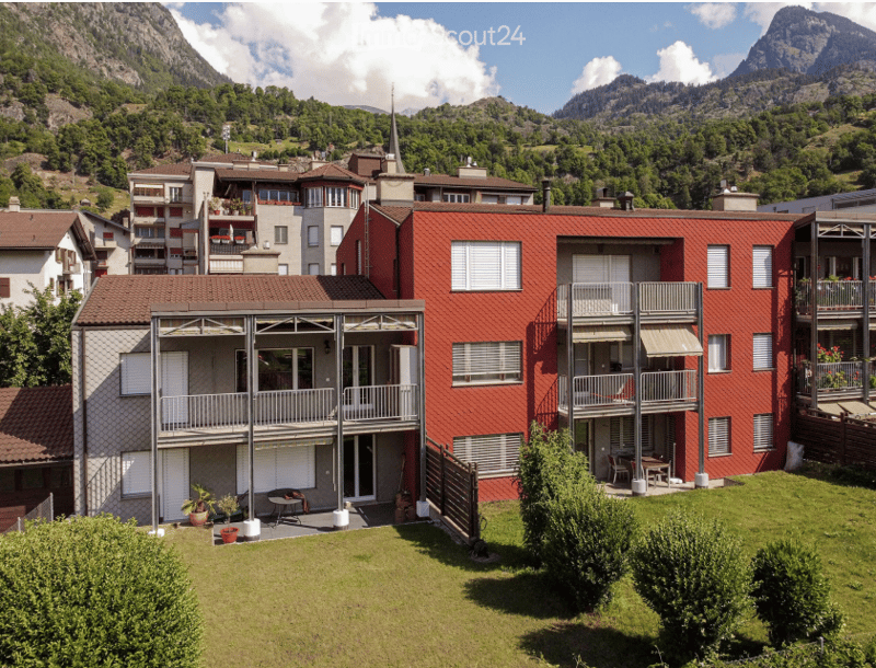Red brick apartment building with multiple balconies and a well-maintained garden area, set against a backdrop of mountains and trees.