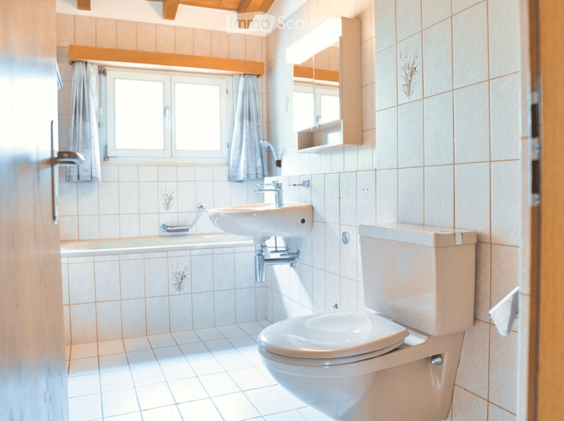Bright bathroom with tiled walls and floor, featuring a sink, toilet, bathtub, and a mirror above the sink. Light-colored curtains cover the window.