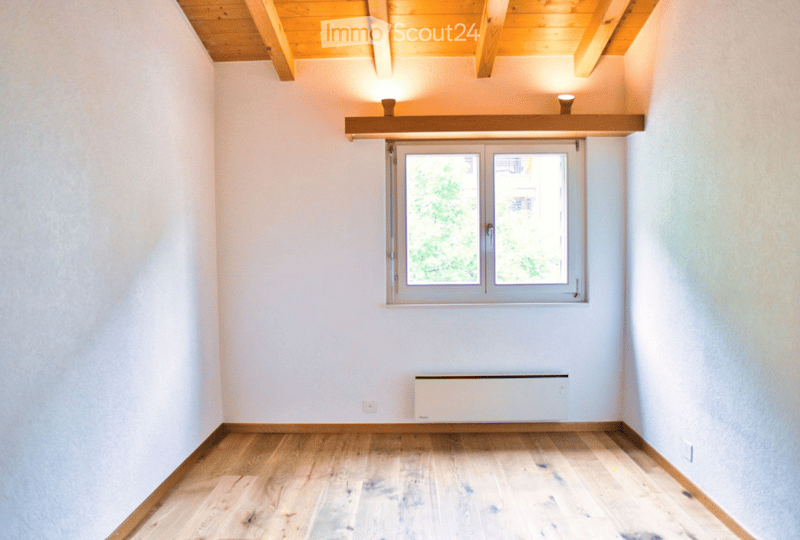 Empty room with wooden flooring, white walls, and a large window with a view of greenery outside. The room features a simple wooden ceiling and a radiator on the wall.