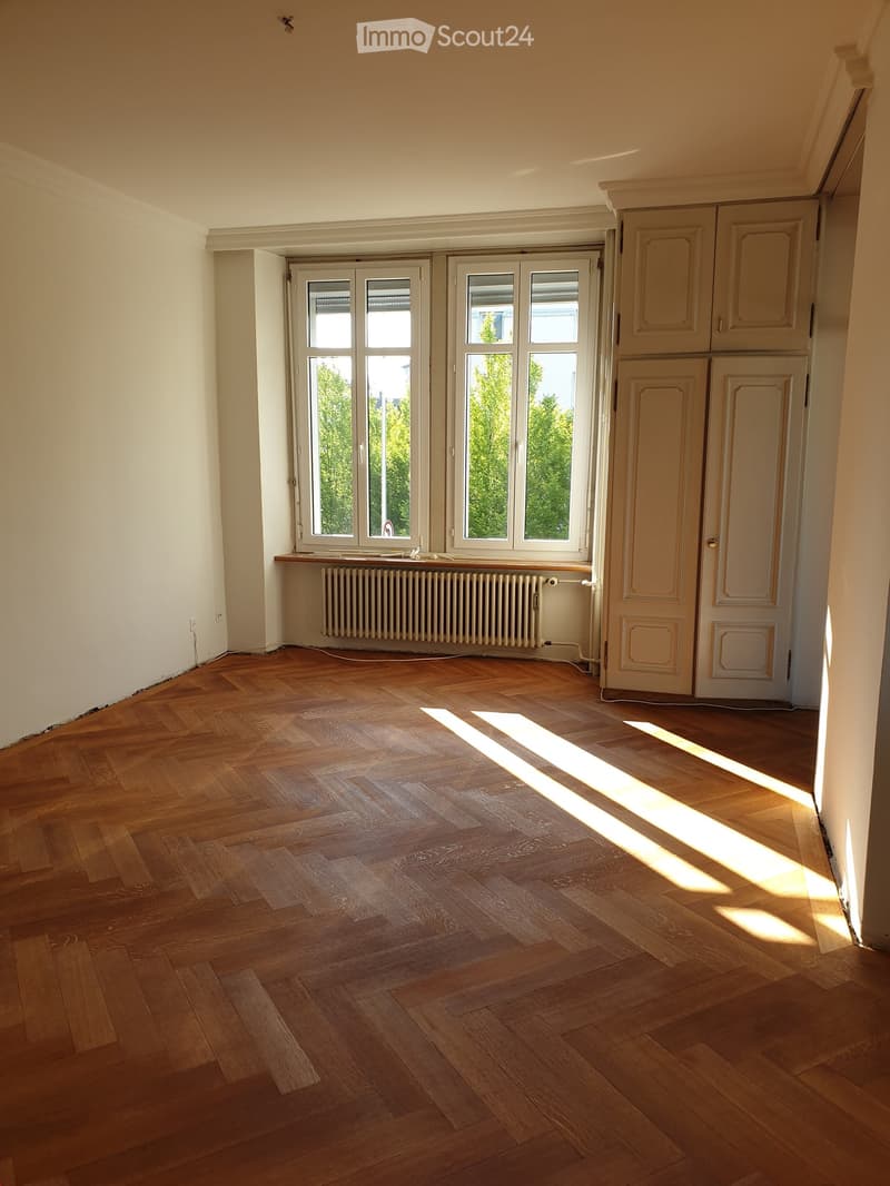Empty room with wooden parquet flooring, large windows with white frames, radiator, and built-in cabinets.