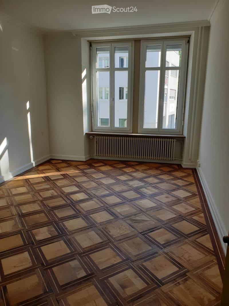 Empty room with wooden floor and large windows, featuring a radiator under the windows and light-colored walls.
