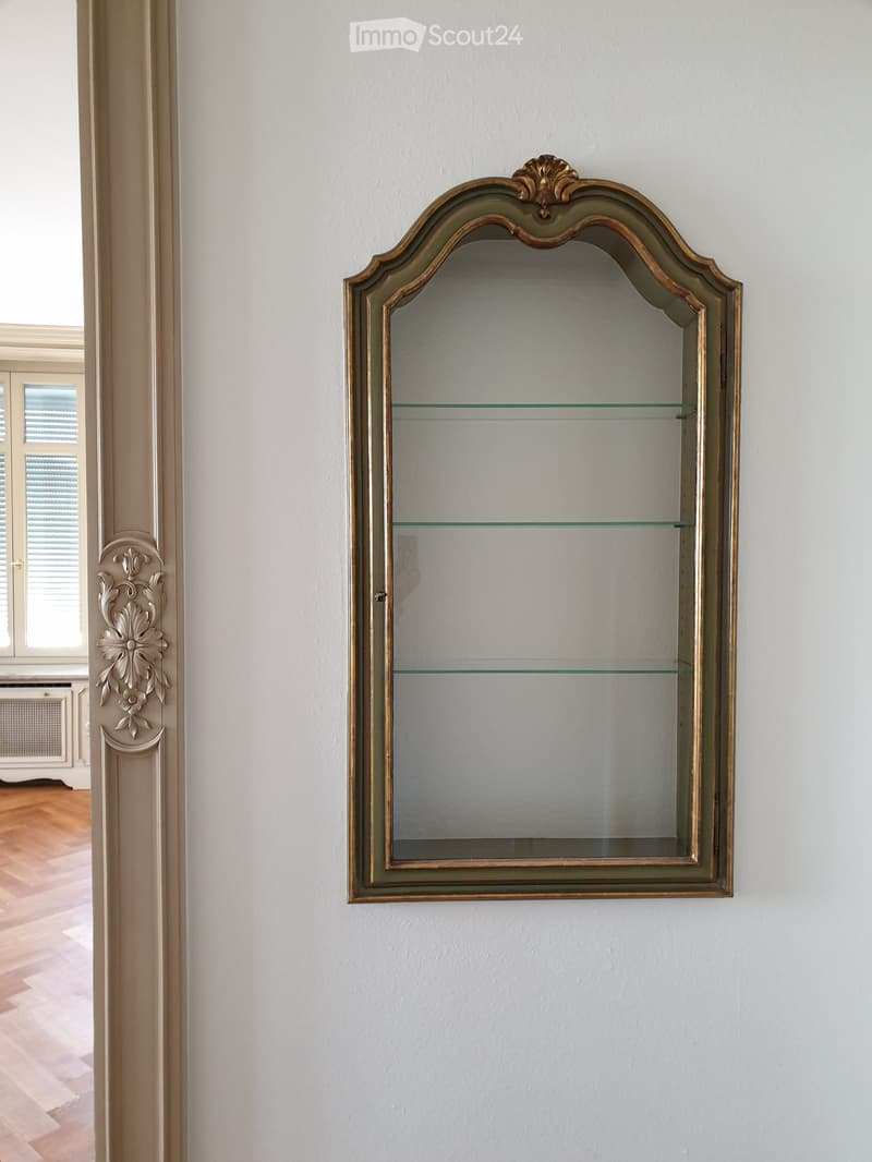 A wall-mounted cabinet with glass shelves and ornate gold detailing, positioned in a well-lit room with light-colored walls and wooden flooring.