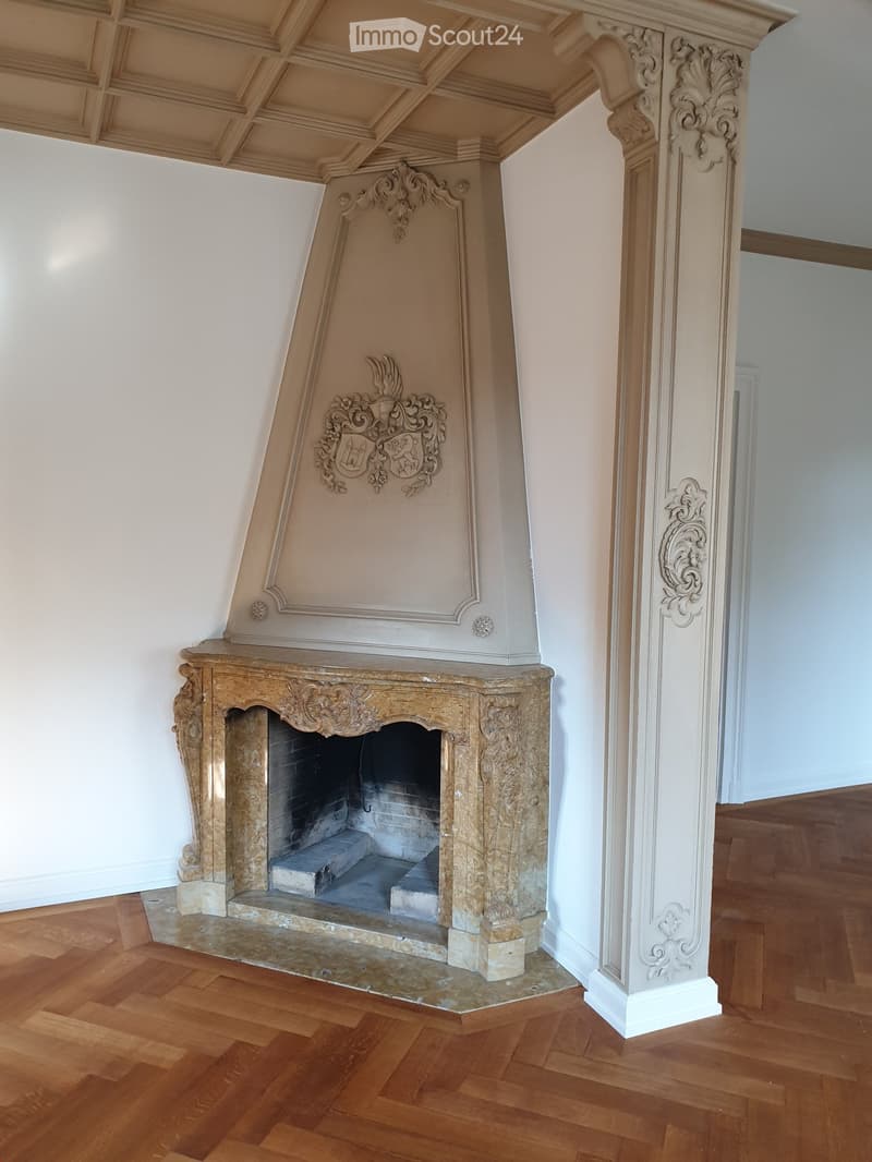 Elegant fireplace with ornate carvings and a decorative crest above, set against white walls and a polished wooden floor.
