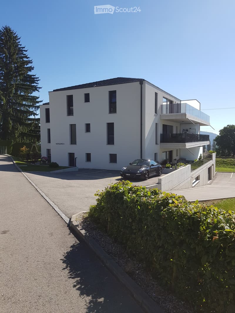 3-story apartment building with a gray exterior, large windows, and a balcony. There is a parking area in front of the building with a car parked. The building is surrounded by trees and landscaping.