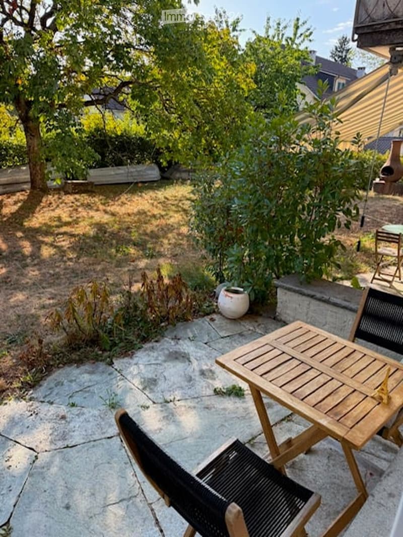 Garden with a stone patio, wooden table, and chairs, surrounded by trees and plants.
