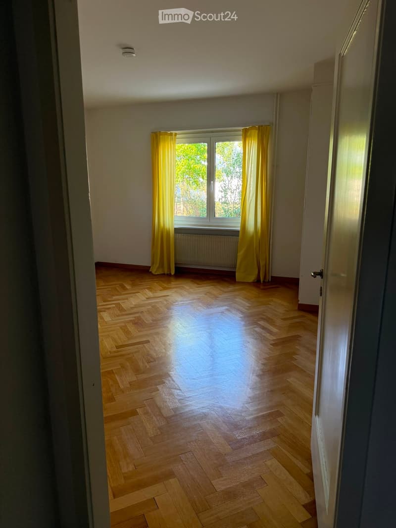Empty room with wooden herringbone flooring, large window with yellow curtains, radiator, and white walls.