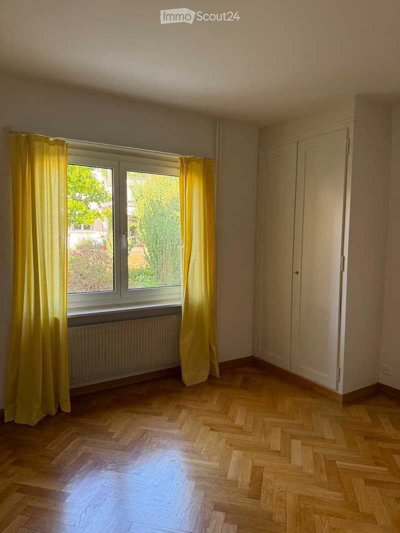 Empty room with wooden herringbone flooring, large window with yellow curtains, radiator, and built-in closet.