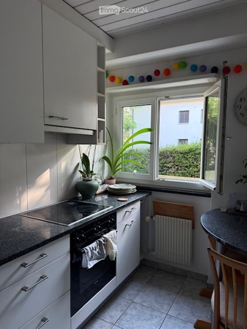 Modern kitchen with white cabinets, induction stove, oven, and a small dining area. Features a large window with a view of greenery outside.
