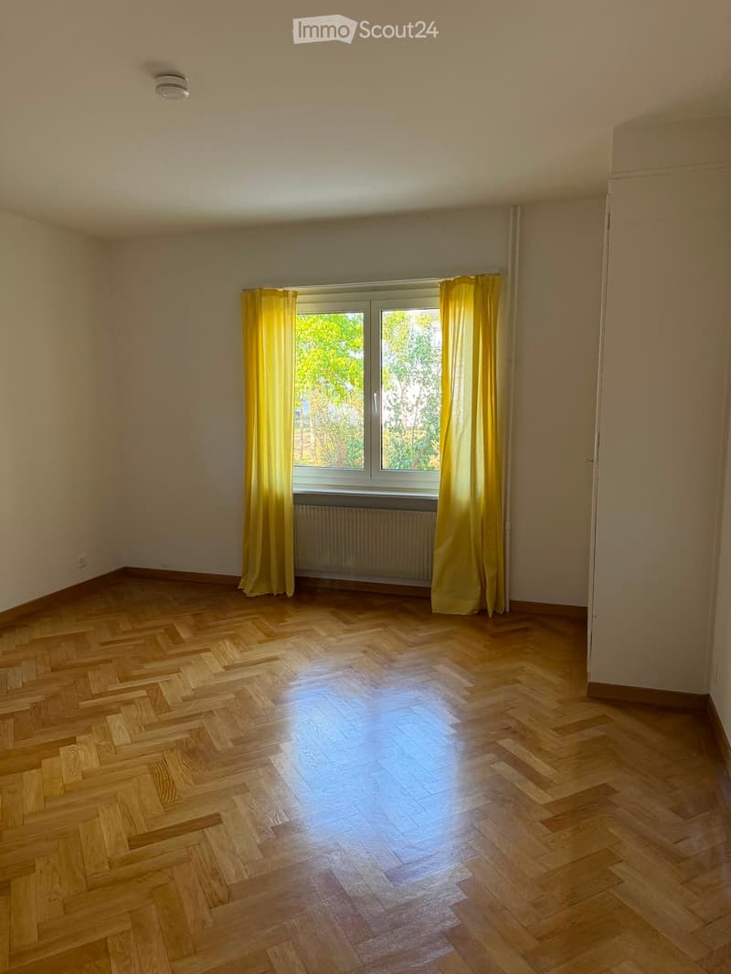 Empty room with wooden herringbone flooring, large window with yellow curtains, radiator, and white walls.