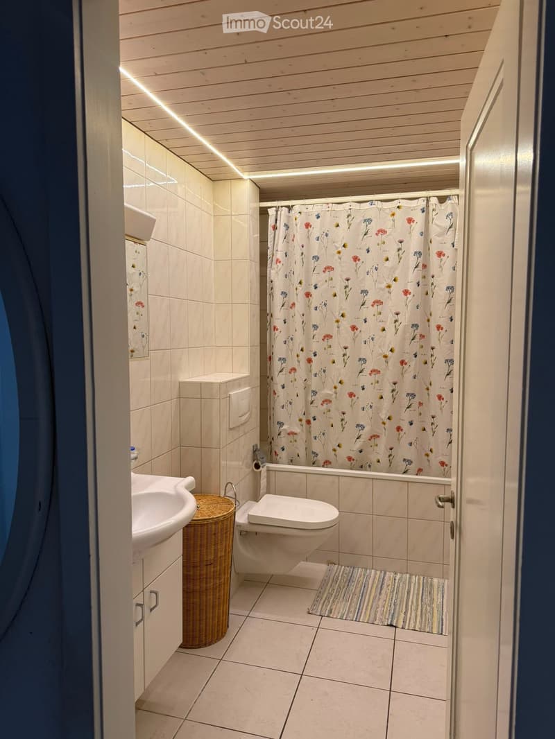 Modern bathroom with white tiles, a sink, toilet, bathtub with floral shower curtain, wicker basket, and a striped bath mat.