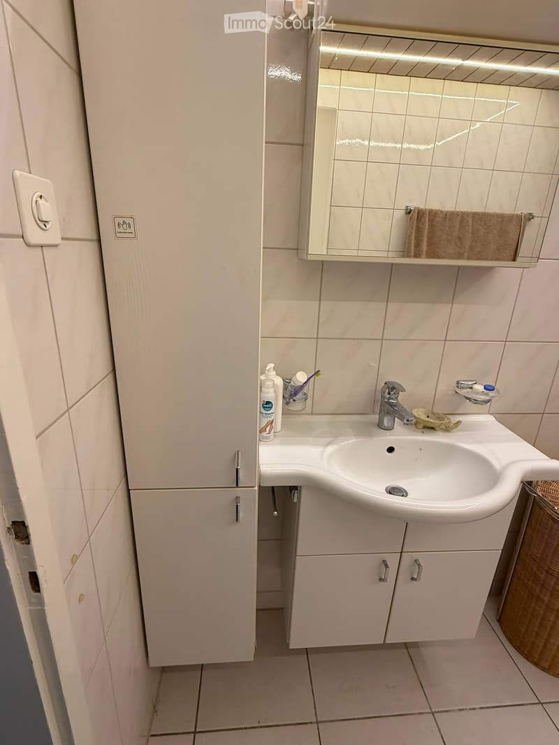 Modern bathroom with a white sink, mirror, cabinet, and various toiletries including a toothbrush, toothpaste, and a bottle. The walls are tiled, and the floor is also tiled.