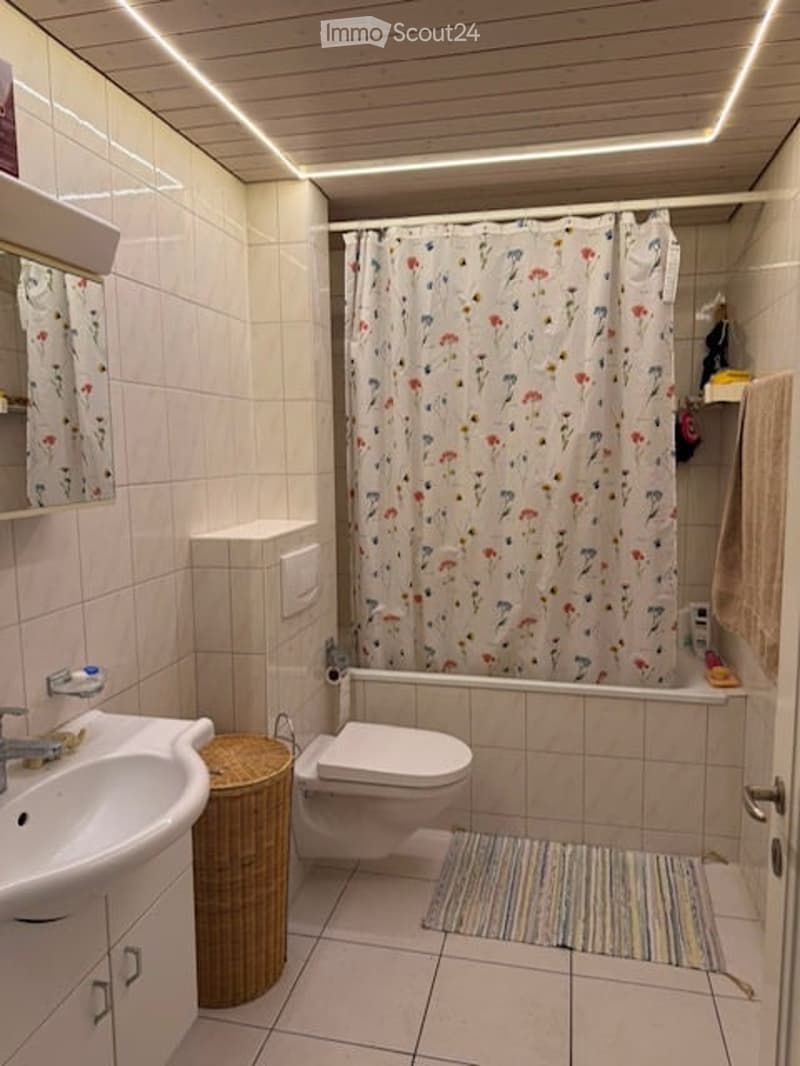 Modern bathroom with white tiles, a sink, toilet, bathtub with a floral shower curtain, a mirror, a wicker basket, and a colorful rug.