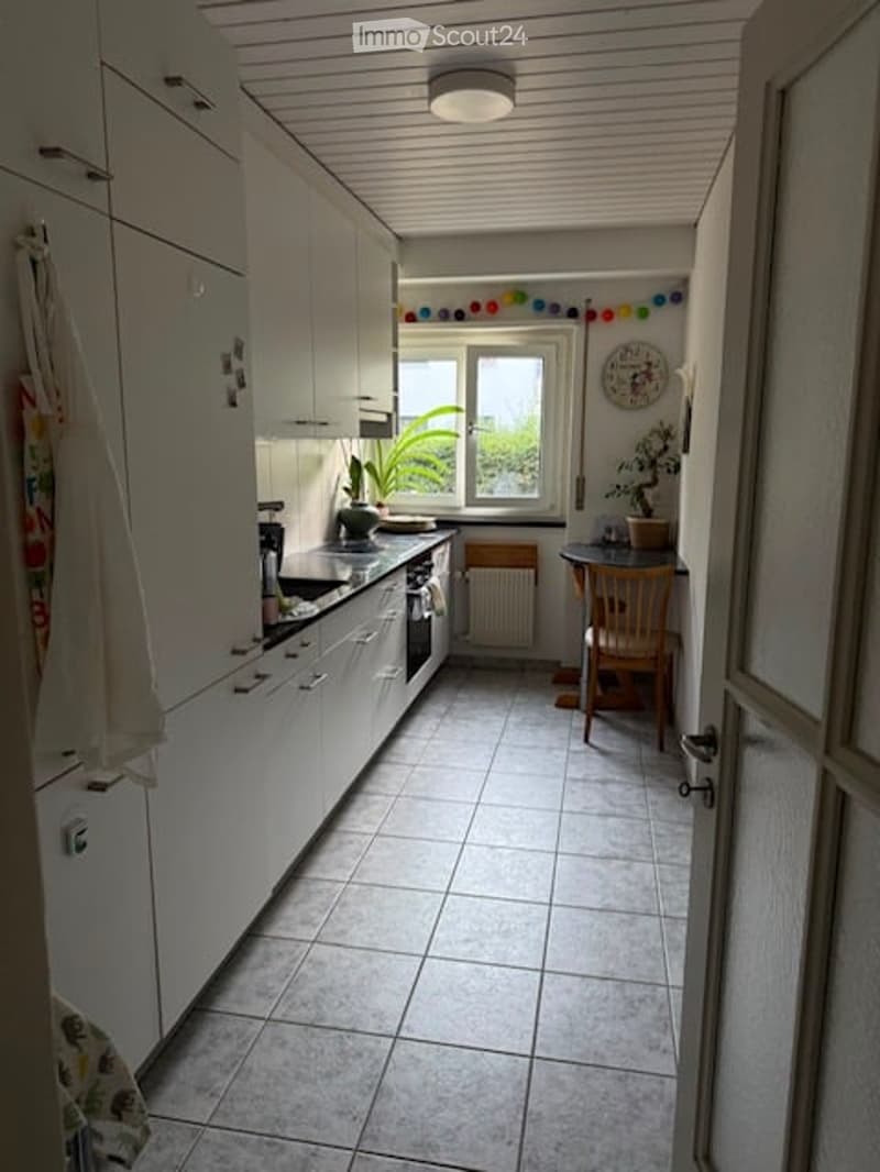 Bright kitchen with white cabinets, tiled floor, and a window overlooking a view. Includes a refrigerator, oven, sink, coffee maker, small table with a chair, and a clock on the wall.