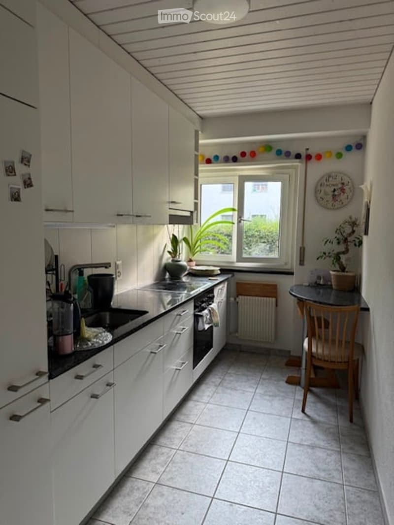 Modern kitchen with white cabinets, stainless steel appliances, and a small dining area with a chair and table. A window offers natural light and a view of greenery outside.