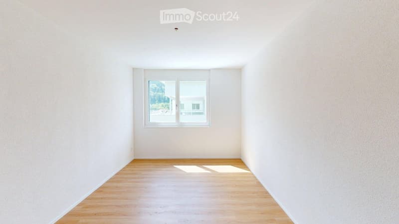 Empty room with light wooden flooring and white walls, featuring a large window with a view of greenery outside.
