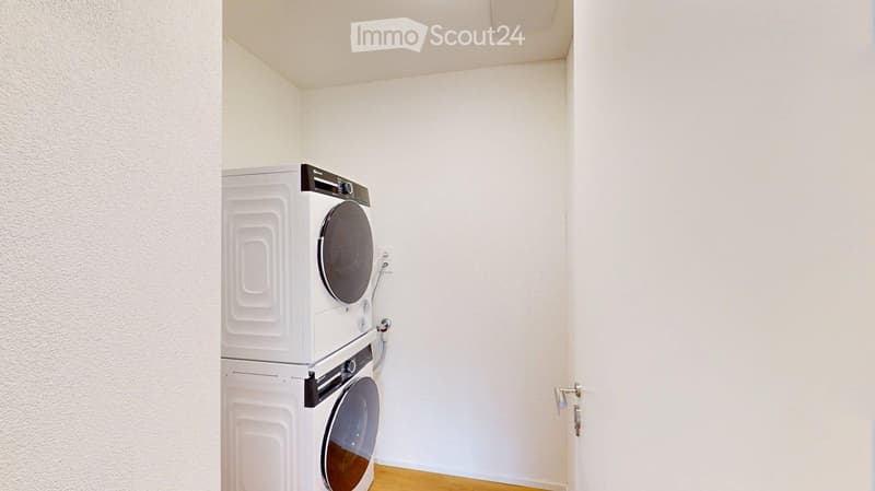 A small, clean room with a white washing machine and dryer stacked vertically against a white wall. The floor is light wood, and the walls are white.