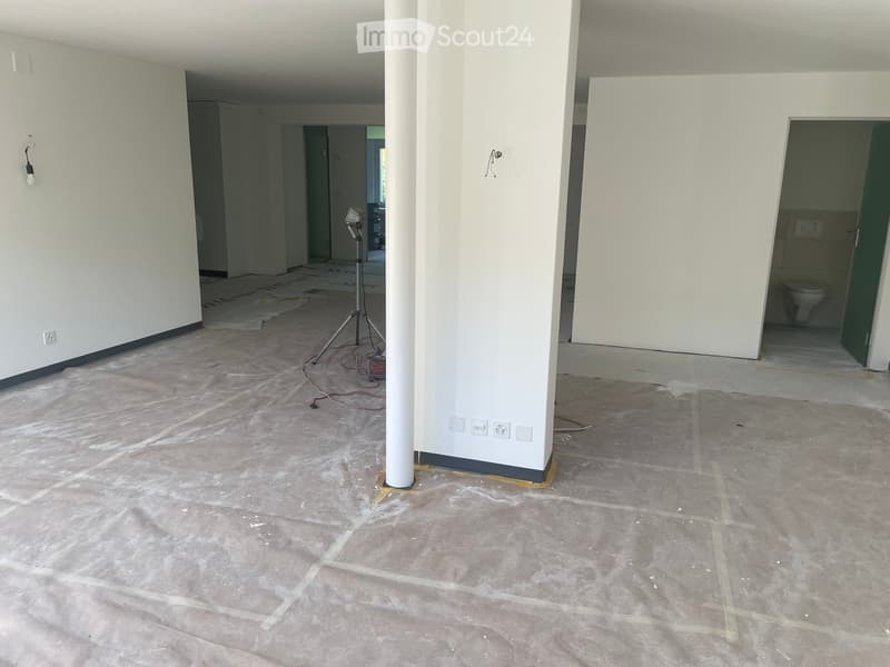 Empty room with unfinished concrete floor and white walls, featuring a central column and visible electrical outlets. A tripod and wires are present, and a partially visible bathroom with a toilet is seen through an open doorway.