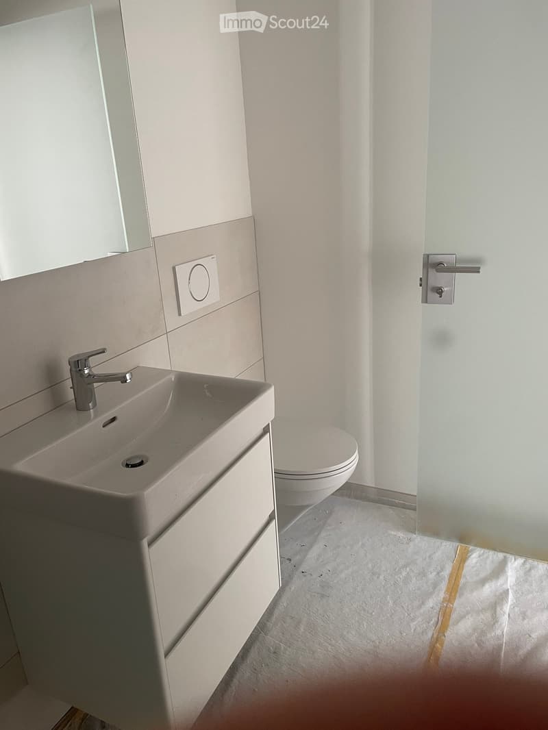 Modern bathroom with a white sink, chrome faucet, and toilet. Light-colored tiles and a glass shower enclosure are visible.