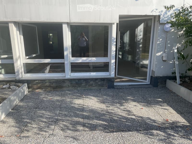 Modern building entrance with large glass doors and windows, raised ground floor, small garden area with rocks and plants.