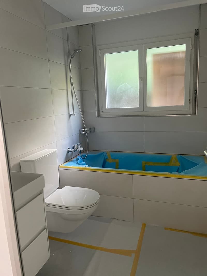 Modern bathroom with a toilet, bathtub, and showerhead. The walls are tiled in light grey, and the floor is covered with a protective blue sheet.