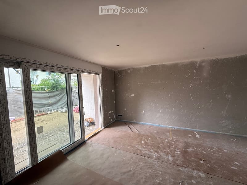Empty room with unfinished walls and floor, large sliding glass door leading outside, visible construction materials and measurements on the left side.