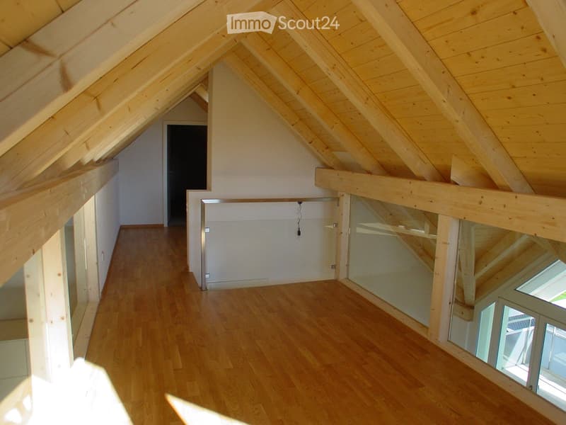 An empty attic space with wooden beams, wooden flooring, and exposed wooden ceiling beams. The room has large windows and a doorway leading to another area.