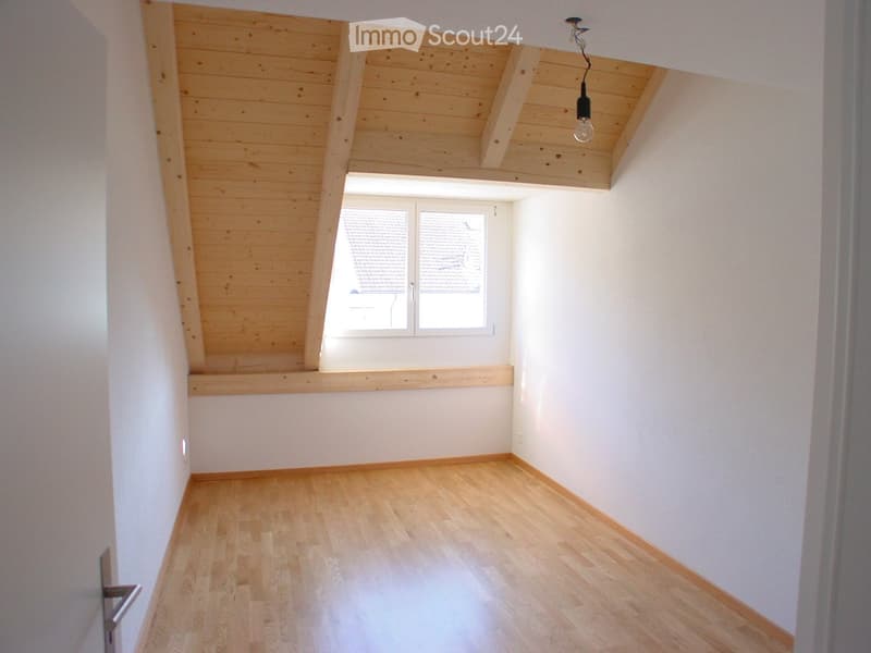 Empty attic room with wooden ceiling, wooden floor, and large window with blinds.