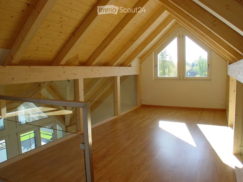 Bright, empty attic space with wooden beams, wooden flooring, large windows, and a glass railing overlooking a lower level.