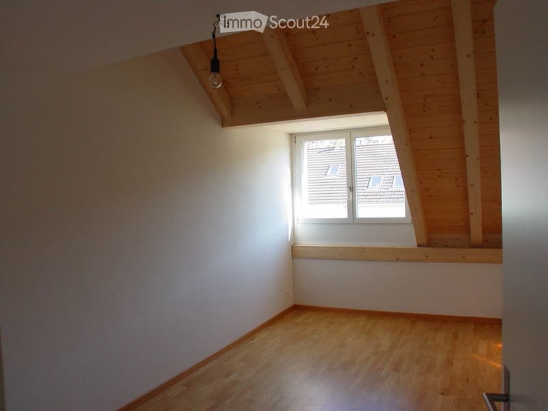 Empty room with wooden flooring, white walls, and a skylight window. The ceiling has exposed wooden beams and a hanging light fixture.